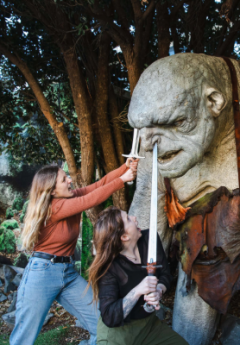 Two women posing with swords against a troll statue.