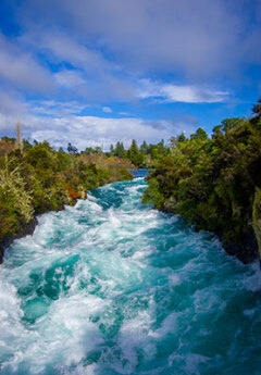 Rushing turquoise waters of Huka Falls surrounded by lush greenery under a partly cloudy sky.