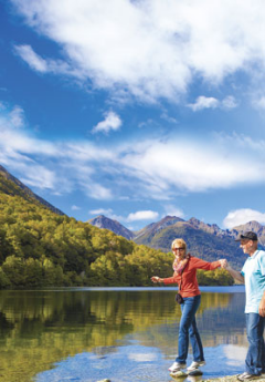 A woman and man walk along the edge of a calm lake under a sunny, blue sky.