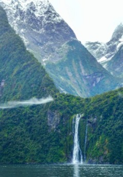 A waterfall plunges into the water between steep, green cliffs and snowy mountains.