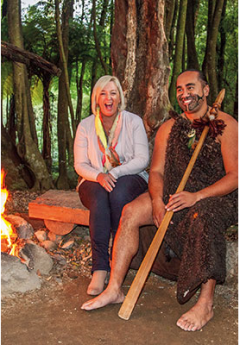 A tourist smiles next to a Māori warrior in a cloak by a campfire in a forest.