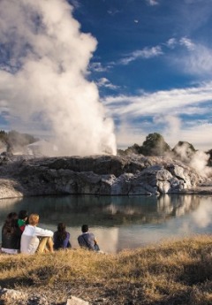 Visitors watch a powerful geyser erupting near a steaming thermal pool.