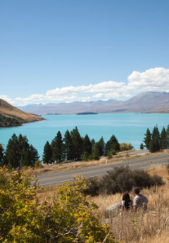 A couple sit in the grass overlooking the bright blue waters of Lake Tekapo
