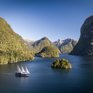 Image of a cruising vessel glides across the still waters of Doubtful Sound, with towering peaks and clear blue sky.