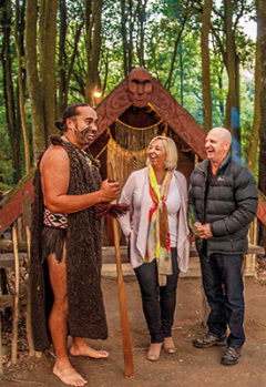 Tourists interact with a man in traditional Māori dress in front of a traditional carved meeting house.