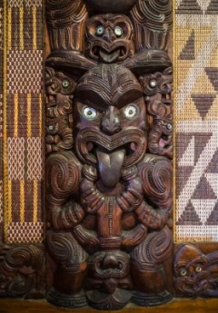 A close-up of an intricate, traditional Māori wood carving with paua shell eyes.
