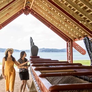 Two women examine a long Māori waka (canoe) under a sheltered structure.