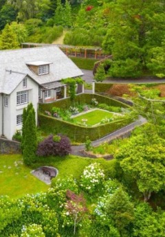 A two-story white house with a manicured garden and hedges, nestled on a green hillside.