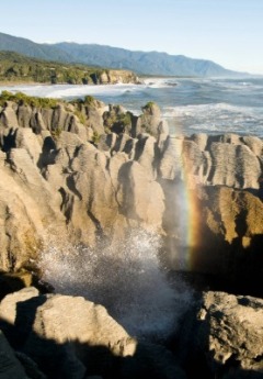 The layered Punakaiki Pancake Rocks spray water creating a rainbow against the coast.