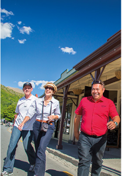 Two tourists and a guide walk down a sunny street past historic buildings with wooden awnings.