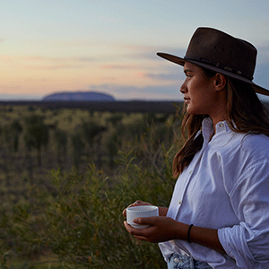 A woman holds a cup of coffee as she gazes over the Uluru landscape at sunrise