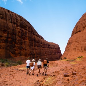 Four people hike toward a narrow gap between massive red rock formations.