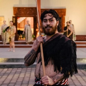A Māori man in traditional dress smiles while holding a wooden taiaha.