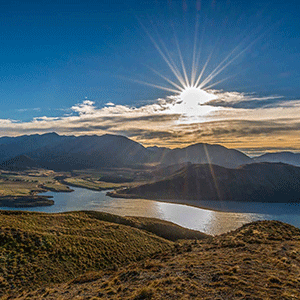 A view of Lake Wanaka from above at sunset, the lake and rolling hills and mountains tinged golden from the light.
