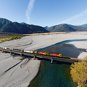 The TranzAlpine Train crosses a river on a raised bridge with mountains in the background.