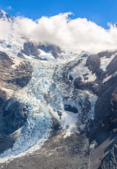 Dramatic landscape of Fox Glacier, with jagged grey stone, icy blue glacier, and clouds.