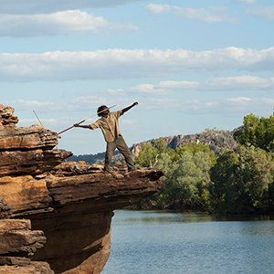 A local Aboriginal guide is lunging backwards with a spear in his hand, standing on a overhanging rockface above the East Alligator River in Kakadu National Park.