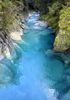 A gorge, viewed from the top down, flanked on both sides by grey rock walls, lined with lush greenery. The water is crystal clear and a vibrant shade of blue.