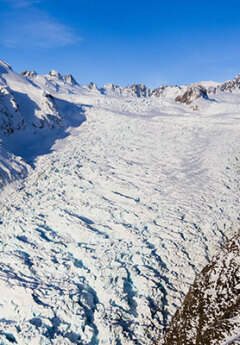 An aerial shot of Fox Glacier, a wide expanse of ice and snow, hugged in on all sides by mountain peaks, a clear blue sky in the background.