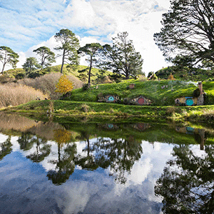 A View of Hobbiton, with round, brightly coloured doors to Hobbit homes set into a rolling greemm hill facing a calm river on a sunny day.