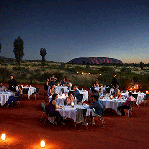 Guests dine under outback skies as the sun sets beyond Uluru in the background