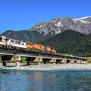 The TranzAlpine Scenic Train travels over a bridge over a braided river, with mountains in the background.