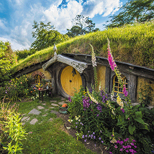 A Hobbit House with a large, round, yellow door is nestled into a hill with green grass and wildflowers.