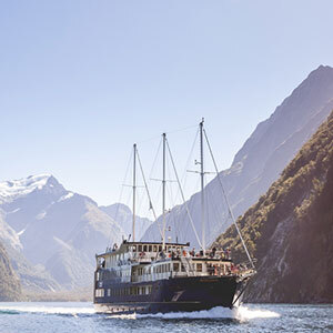 A cruising vessel is in Milford sound, surrounded by sky-high mountain peaks, some with snow, and a clear blue sky.
