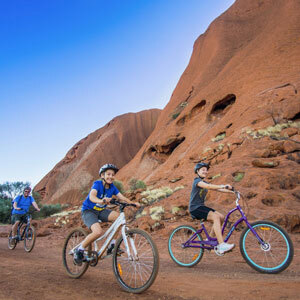 A close up image of two children riding their bicyles around the giant red rock, Uluru.
