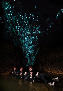 Three people sit on a boat on the water in the darkness with blue glowing worms filling the walls