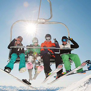A groupd of four are on a chair lift, viewed from slightly below and in front, three skiiers and one snowboarder on a blue sky sunny day.