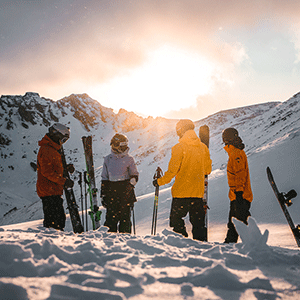 A group of four people are on a snowy mountain with an assortment of skis and snowboards at sunrise, the sky golden and blue.