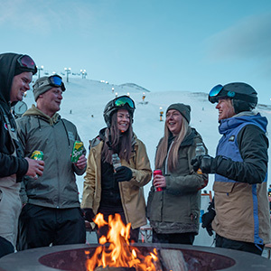 A group of 5 skiers around a bonfire enjoying Apres with the lights of the Coronet Express chairlift and white snow in the background