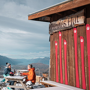 Two skiers enjoy a break at Heidi's Hut mountain bar on Coronet Peak. The hut is covered in skies and there are mountains in the background