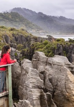 A woman on a boardwalk views the jagged, layered Punakaiki Pancake Rocks and coast.