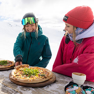 Two females in ski clothing share a pizza at Cardrona