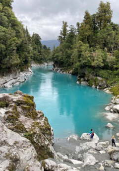 A bright blue lake disapears down a gorge and people climb on the silver rocks surrounding the lake