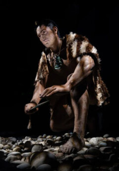 A Maori man crouches on stones looking ahead. His body fades into the dramatic shadows cast around him