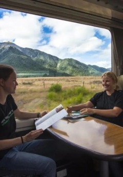 Two women smile while relaxing on a train with scenic mountain views outside.