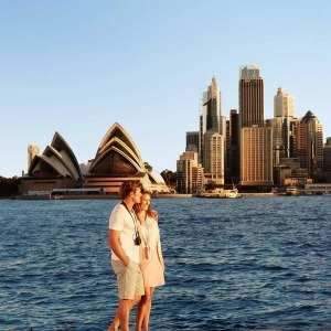 A couple stands by the sea with the Sydney Opera House and city skyline in the background.