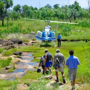 Tourists walk toward a waiting helicopter in a lush, grassy landscape.