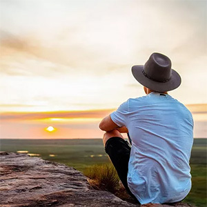 Man on cliff edge watches sunset over plains.