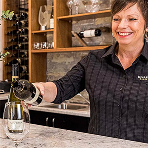 A smiling woman pours wine at a tasting bar.