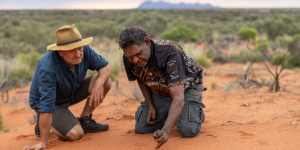 Two men crouch in red sand examining animal tracks