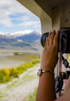 Person photographing mountains from TranzAlpine train outdoor viewing carriage.