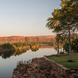 Calm river flows past red cliffs and green palms at sunset.