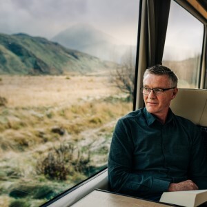 A man sits by a train window looking out at a grassy, mountainous landscape.