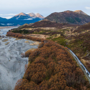 A scenic train travels alongside a braided river with mountains in the background.