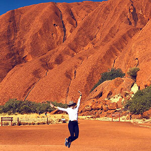 A woman in a whit shirt and black pants star jumps in front of the bright red dirt of Uluru