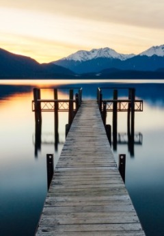 A long exposure of a wooden pier on a calm lake with a sunset and snowy mountains.